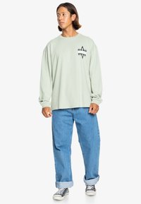 Light green long-sleeve shirt with black graphic design on the chest, paired with blue relaxed-fit jeans and black and white sneakers.