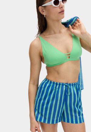 Kobieta w białych okularach przeciwsłonecznych, zielonym topie bikini i niebieskich szortach z zielonymi pionowymi paskami, trzyma ręcznik na ramieniu.