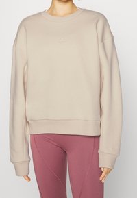 Personne portant un sweatshirt beige Adidas avec le texte "ALL SZN" sur la manche et un legging taille haute mauve, se tenant devant un fond neutre.