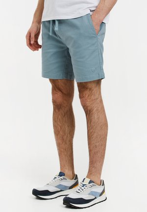 Pantalones cortos de algodón azul claro con bolsillos laterales y cintura ajustable con cordón. Llevados con zapatillas deportivas multicolores con suelas prominentes y tela de malla.