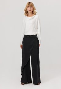 Biała, długa bluzka z dekoltem w łódkę, połączona z czarnymi spodniami typu wide-leg z zasłonięciem, które mają boczny detal w postaci pierścienia i gładką teksturę.