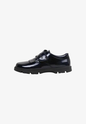 Zapatos de charol negro con una sola tira de Velcro y un detalle decorativo de borlas, que cuentan con una suela de goma negra gruesa y una punta redondeada.