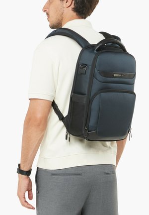 Man wearing white shirt and gray pants with dark blue structured backpack featuring multiple zippered compartments.
