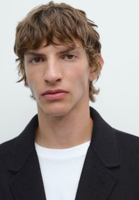 Portrait of a person with tousled brown hair, wearing a black jacket over a white shirt, set against a neutral background.