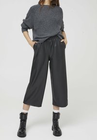 Schwarze, weite Hosen aus Kunstleder mit elastischem Bund, kombiniert mit einem grauen, gerippten Pullover. Schwarze Schnür-Stiefeletten mit gepolsterten Akzenten.