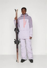 Persona che indossa un abbigliamento da sci di un viola chiaro e stivali da sci neri, in piedi mentre tiene un paio di sci in posizione verticale contro uno sfondo semplice.