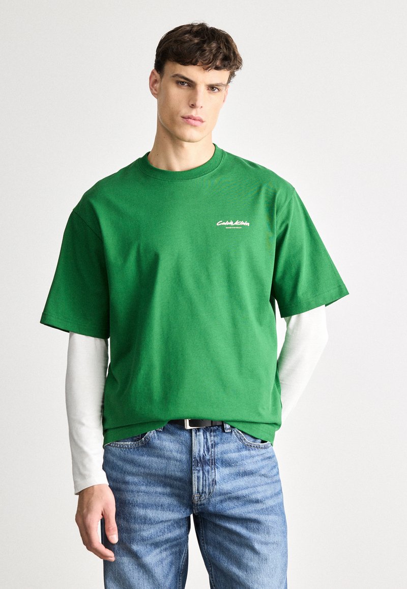 T-shirt de algodão verde com mangas curtas, apresentando um decote redondo e um logotipo branco no peito, combinado com jeans de ganga azul claro.