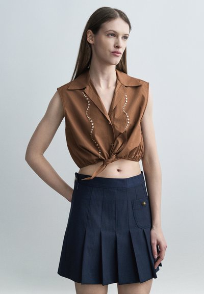 Jeune femme portant un crop top marron sans manches noué avec des clous et une jupe plissée bleu marine avec une poche latérale, sur un fond uni.