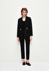 Svart skräddarsydd kostym med dubbelknäppt kavaj med krämfärgade knappar, i kombination med slim-fit byxor och svarta läderloafers.