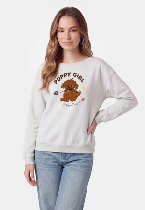 Kvinde iført hvid sweatshirt med brun hvalp illustration og tekst "Puppy Girl I Love Puppies,", kombineret med lyseblå jeans og blå øreringe i bøjle.
