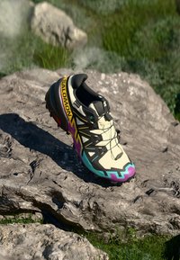 Shoe atlético de cor clara com detalhes pretos e coloridos. Apresenta uma sola texturizada, padrão único e um logotipo da marca em destaque na lateral.