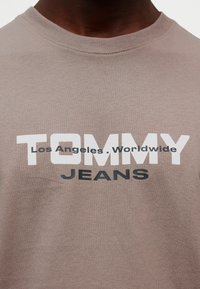 Καφέ βαμβακερό t-shirt με στρογγυλή λαιμόκοψη και μεγάλο τυπωμένο κείμενο: "TOMMY" σε λευκό, "Los Angeles . Worldwide" σε μαύρο και "JEANS" σε μπλε.