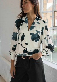 Vrouw in een witte blouse met grote zwarte bloemenprints en zwarte broek, staande bij een raam met één hand in de zak.