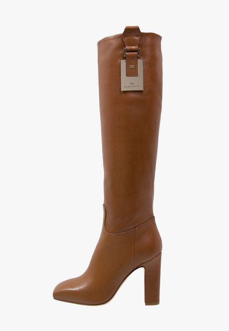 Brązowy kozak sięgający do kolan ze skóry, z kwadratowym noskiem, wysokim szerokim obcasem oraz metalową ozdobą u góry. Gładka tekstura i dopasowany fason.