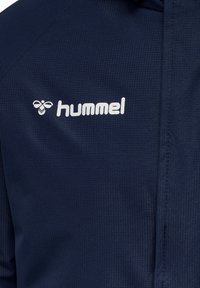 Mørkeblå tekstureret stof med hvidt hummel-logo og bi-ikon på brystområdet af en jakke eller sportstøj.