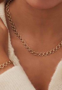 Collier chaîne en or et bracelet assorti avec un design de cercles liés portés sur une peau mate, associés à un vêtement tricoté blanc doux.