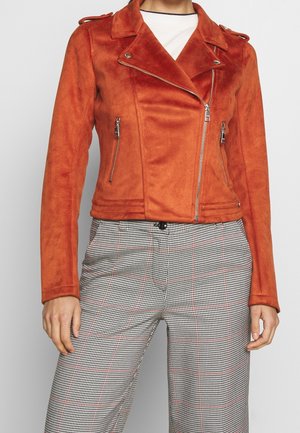 Kobieta w kurtce motocyklowej zamszowej w kolorze rdzawym pomarańczowym z srebrnymi zamkami błyskawicznymi oraz w czarno-białe spodnie w kratę z subtelnymi czerwonymi liniami.