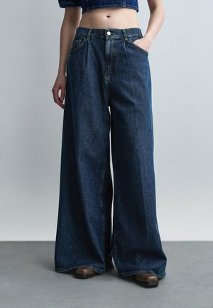 Osoba nosi tmavomodré široké rifle s rukou v vrecku, doplnené hnedými topánkami a skráteným denimovým topom.