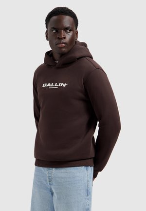 Jonge man in een donkerbruine hoodie met de tekst "BALLIN'", zijwaarts kijkend, met lichtblauwe spijkerbroek tegen een effen lichte achtergrond.