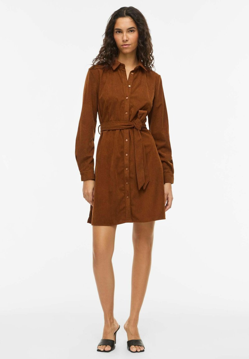 Robe en velours côtelé marron avec manches longues, boutonnée sur le devant et détail de taille nouée. Comprend un col et un ourlet arrivant au genou. Sandales noires style mule.