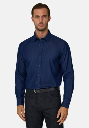 Uomo che indossa una camicia blu scuro a maniche lunghe con bottoni infilata in pantaloni neri con cintura nera, su uno sfondo semplice.