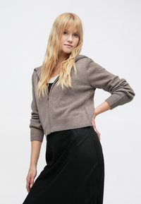 Hoodie raccourci à fermeture éclair en tissu tricoté taupe doux. Doté d'une capuche et de poignets côtelés, associé à une jupe en satin noire.