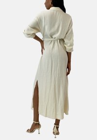 Vestito in tessuto bianco stropicciato con maniche lunghe, vita sottile e spacchi laterali alti, abbinato a scarpe con tacco alto e cinturini leggeri.
