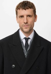 Schwarzer zweireihiger Wollmantel mit Reverskragen, getragen über einem grauen Weste und weißem Hemd, ergänzt durch eine schwarze Krawatte.