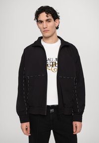 Schwarze Zip-Jacke mit weißen Nähten, Stehkragen und elastischen Bündchen, kombiniert mit einem weißen Grafik-T-Shirt und dunkelblauen Jeans.
