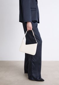 Borsa in pelle bianca con un design geometrico, dotata di una maniglia sottile e dettagli in catena argentata, tenuta da una persona in abbigliamento scuro.