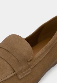 Mocassins en daim marron avec un design couturé, une texture lisse et une semelle en caoutchouc ornée d'une bande décorative sur le dessus.