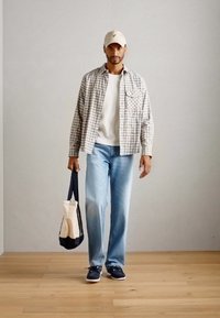 Mann iført beige caps, rutete skjorte over hvit t-skjorte, lyseblå jeans, marineblå joggesko, holder en beige og svart tote bag, står på tregulv.
