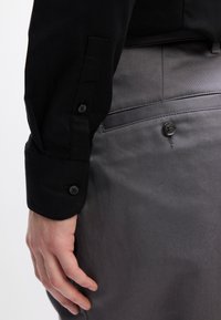 Zwarte gestructureerde shirt met knoopsluiting bij de manchetten, gecombineerd met grijze gladde broek met twee knopen op de achterzak.