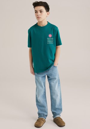 Teal Baumwoll-T-Shirt mit einer pinken Sonne und wavy Design, kombiniert mit hellblauen Jeans und braunen Mokassins.