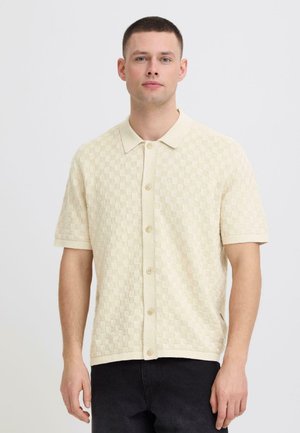 Mann iført beige kortermet rutete strikket skjorte med knapp og krage, stående mot en ensfarget hvit bakgrunn.