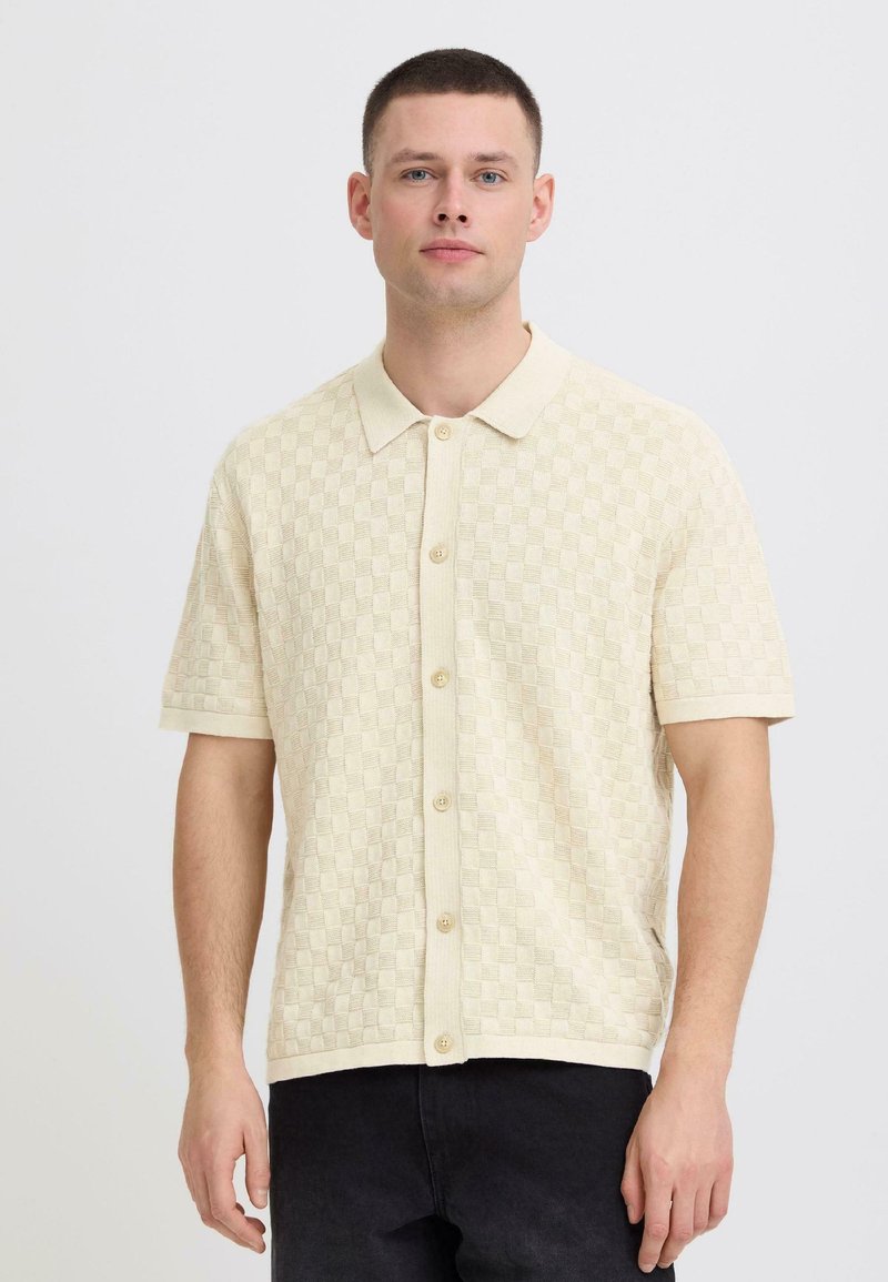 Homme portant une chemise en tricot à manches courtes beige à carreaux avec col et boutons, debout devant un fond blanc uni.