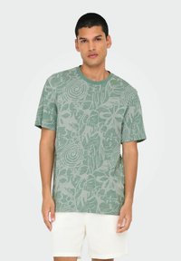 Camiseta de algodón verde con un patrón floral texturizado. Mangas cortas y escote redondo. Usada con pantalones cortos beige.