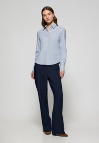 Camicia azzurro chiaro con colletto, maniche lunghe e un sottile motivo a righe, abbinata a pantaloni a gamba larga blu scuro e scarpe marroni.