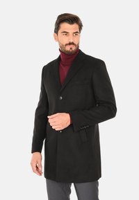 Cappotto di lana nera con chiusura a un bottone, revers a scensione e tasca frontale, indossato su un dolcevita bordeaux. Vestibilità su misura.