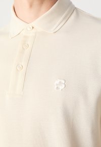 Polo beige à manches courtes avec trois boutons et petit logo blanc brodé sur la poitrine gauche, porté par une personne.