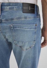 Hellblaue Denim-Jeans mit einer Gesäßtasche und einem schwarzen Label mit der Prägung "MAVI 911 ULTRA MOVE". Strukturierter Stoff mit leichter Ausbleichung.