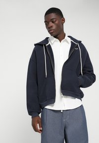 Ναυτικό μπλε φούτερ με φερμουάρ και κορδόνια, μαλακό ύφασμα, μπροστινές τσέπες, φορεμένο πάνω από λευκό πουκάμισο με κουμπιά, συνδυασμένο με ανοιχτόχρωμο τζιν παντελόνι.