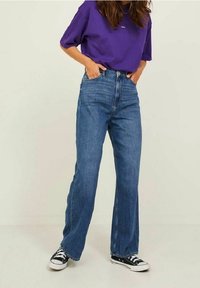 Žena v volných modrých džínách, černo-bílých teniskách a fialovém velkém tričku, která stojí s rukama v kapsách na jednotném pozadí.