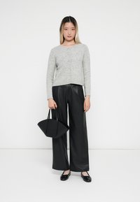 Cardigan en tricot gris clair avec fermeture à boutons, porté sur un pantalon large en simili cuir noir et des chaussures plates noires, tenant un sac noir.
