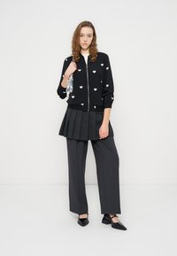 Νεαρή γυναίκα που φοράει μαύρο πουλόβερ με φερμουάρ και λευκές καρδιές, πλισέ φούστα πάνω από φαρδιά γκρι παντελόνια, μαύρα μυτερά παπούτσια και κρατά μια ασημένια τσάντα.