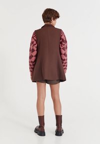 Brown sleeveless outerwear over a pink patterned sweater, paired with dark shorts and brown ankle boots. Smooth fabric and structured design.