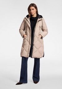 Béžový prošívaný péřový kabát s černou kapucí, se zipovými kapsami a střední délkou. Nošený přes tmavé rozšířené džíny.