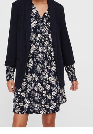 Mujer con un vestido azul marino con flores blancas y rosa claro, combinado con un blazer negro con las mangas remangadas.