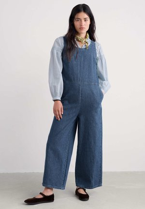 Denim overall met een losse pasvorm, voorzien van brede pijpen, een ronde halslijn en zijzakken. Gecombineerd met een lichtblauw blouse en donkere schoenen.
