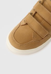 Pruun nubukist tennisjalats kolmel Velcro rihmal, valge kummitald ja tekstuuriga viimistlus. Õmblused on dekoreeritud servade ümber.